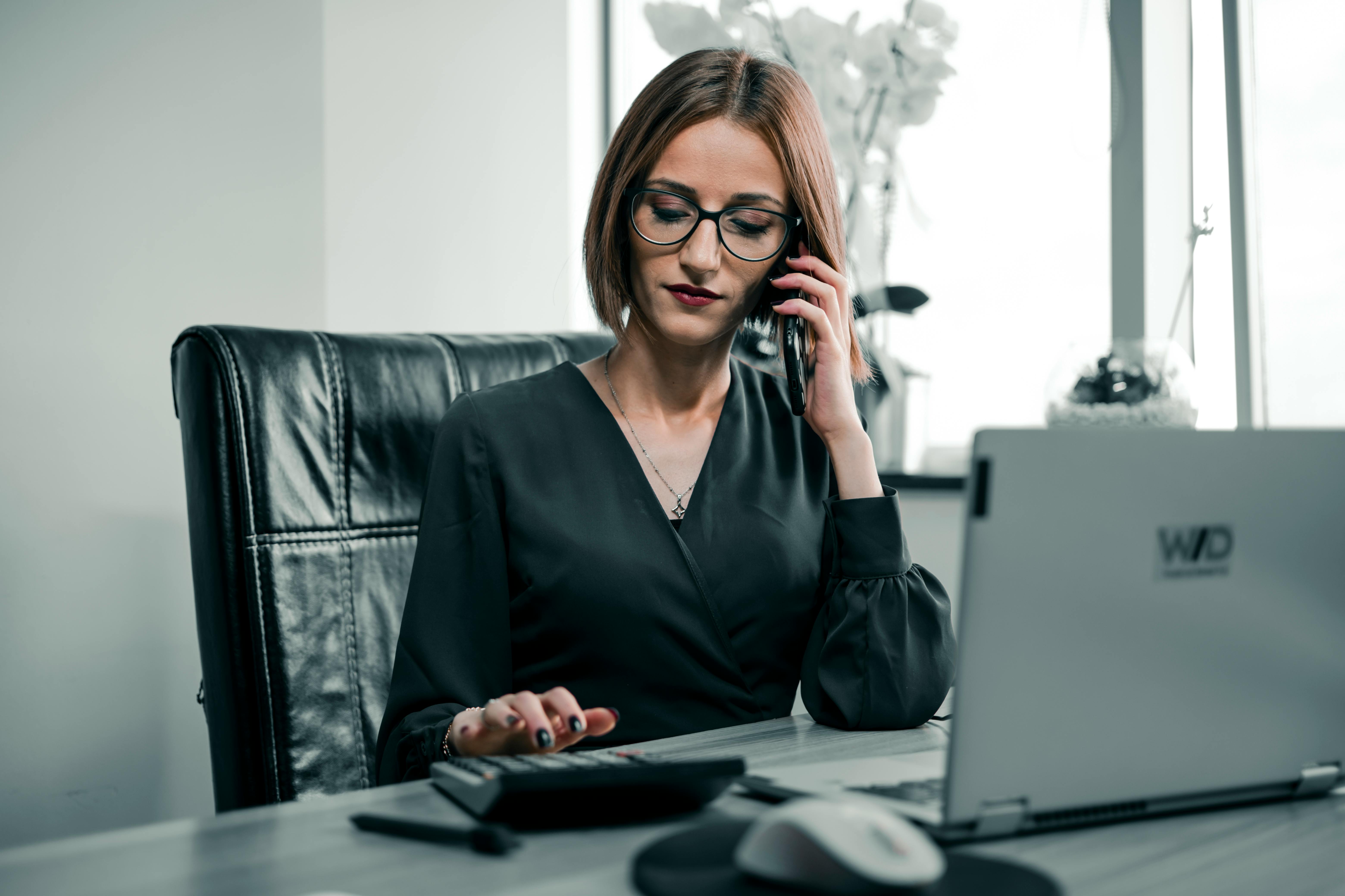 Woman Sitting by Office Desk Taking Call while Using Calculator